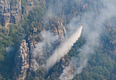 Ein Lastenhubschrauber aus Österreich fliegt mit einem Löschwasser-Außenlastbehälter um einen Waldbrand im Nationalpark Sächsische Schweiz zu löschen.