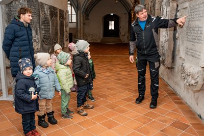 Holger Graf erklärt Kita-Kindern mit Erzieher Jonas Eichhorn die Grabplatten im Kreuzgang.