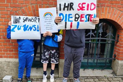 Drei Kinder halten Plakate hoch. Sie wollen erreichen, dass die Assistenten an ihrer Schule bleiben können.
