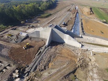 Das Luftbild zeigt das fast fertige, durchlässige Absperrbauwerk sowie den davor und dahinter in Teilen aufgeschütteten Damm von 535 Meter Länge.