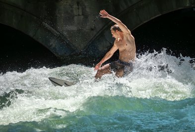 Patrick surft bei sommerlichen Temperaturen mit seinem Board auf der Eisbachwelle im Englischen Garten in München.