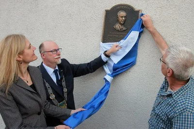 Die Leiterin des Trias-Gymnasiums und der Mittelschule, Cornelia Kiesewetter, Bürgermeister Sandro Bauroth (FDP) und Grundschulleiter Thomas Müller (rechts) enthüllten am Montagabend die Ehrentafel an der Fassade des Elsterberger Schulzentrums, das jetzt Wolfram Markert heißt. 