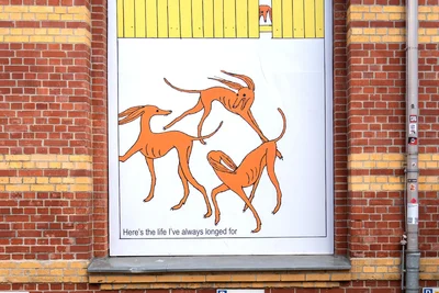 Ein Bild der Comiczeichnerin Anna Haifisch an der Fassade des Wirkbaus neben dem Ausstellungseingang: Hunde tollen frei umher, aber einer schaut isoliert durch die Lücke eines Gartenzauns zu. Ein Bild, das für Einsamkeit, für Ausgrenzung stehen kann.
