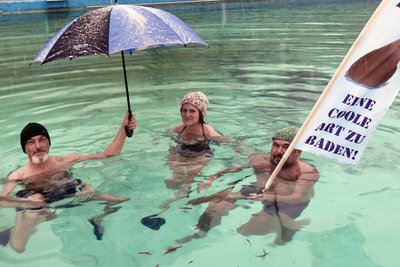 Michael Friedl, rechts mit Motto-Flagge, sowie Ines und Stefan Fuchs beim angestammten Mittwochs-Schwimmtreff im Naturbad Falkenau. Der Regen machts nix, sie haben doch einen Schirm, gegen die Kälte hilft die Mütze.
