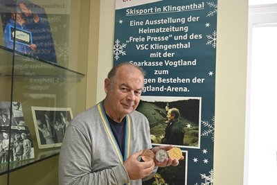Thorald Meisel mit Medaillen von Wettbewerben aus dem Jahr 1967 vor einem Plakat von 2011 zum fünfjährigen Bestehen der Vogtland-Arena.