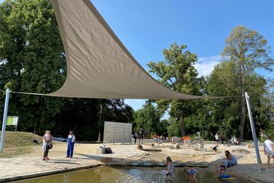 Ein dreieckiges Segeltuch überspannt einen Teil des Wasserspielplatzes und gibt spielenden Kindern Schatten.