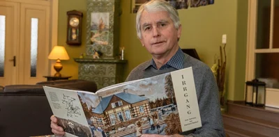 Eberhard Baldauf mit seinem ersten Buch, das sich der Geschichte des Irrgang-Grundstücks und des benachbarten Areals (bis vor kurzem BG-Klinik) widmet. Baldauf sitzt in einer der ehemaligen Irrgang-Gaststuben, die heute sein Wohnzimmer ist. Das Cover-Bild hat er selbst in Öl gemalt. 