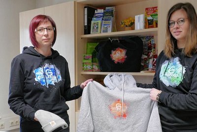 Nancy Müller (links) und Stephanie Wolf präsentieren einige der Kleidungsstücke, die im Zuge der Gelenauer750-Jahr-Feier zu haben sind. Erste Bestellungen liegen der Gemeinde bereits vor.