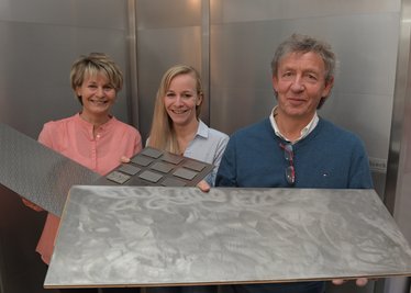 Volker Queck (rechts) zeigt die allererste Edelstahl-Bodenplatte, mit der er zu experimentieren anfing. Seine Frau Katrin (links) hält eines der inzwischen patentierten Paneele, Tochter Josefine präsentiert die zwölf derzeit verfügbaren Oberflächen-Varianten. Im Hintergrund eine Wand aus Queck-Edelstahl-Segmenten.