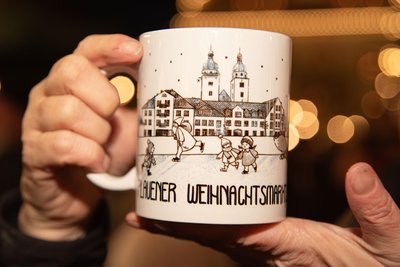 So sieht das neue Weihnachtstipfl für das Jahr 2025 aus. Die Tasse ziert die Johanniskirche, die Eisbahn am Topfmarkt sowie Vater & Sohn und Stadtfüchsin Plauline beim Eislaufen.