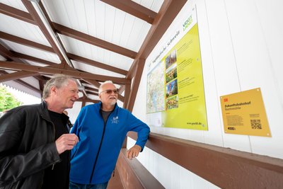 Peter Kilian, leitender Bauüberwacher, und Wanderwegewart Andreas Lehmann betrachten die Infotafel im denkmalgeschützten Wartehäuschen.
