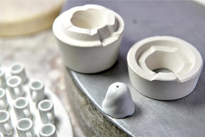 Die Detailaufnahme zeigt das Ober- und das Unterteil einer klassischen Gipsform mit einem fertig gebrannten und glasierten Miniglöckchen. Links im Bild Mini-Glühweinbecher.