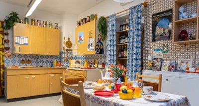 Blick in die Küche mit Sitzecke. Der Tisch ist gedeckt, die Schränke und Regale sind gefüllt - als seien die Bewohner nur kurz außer Haus. 