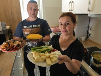 Elena und Alin Cracana aus Rumänien haben in ihrer Wohnung zu Mittag gekocht. Elena bringt einen gemischten Teller mit Käse zum Tisch, Alin einen Slanina-Wurstteller und Màmàligà-Kuchen. 
