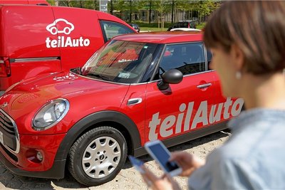 Eine Frau bucht mit einem Smartphone ein Auto des Anbieters Teilauto auf einem Parkplatz. Das Angebot an Fahrzeugen wächst.