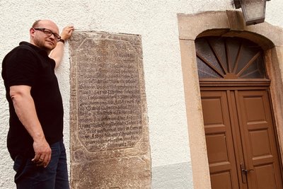 Falk Drechsel an der Auerbacher Kirche, am ältesten Grabstein im Ort. Dieser benennt Georg Viehweger - 1618 geboren, 1690 gestorben -, der fast 52 Jahre im Schuldienst arbeitete.