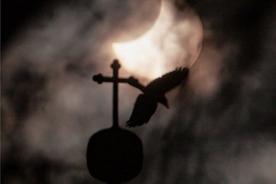 Mystisch: Eins der beiden Turmkreuze der St.Martins-Kirche in Oberlungwitz vor der Sonne, die zur Zeit der größten Bedeckung durch den Mond nur kurzzeitig durch die Wolkendecke schaut. Im Moment des Fotos flog eine Krähe durch das Bild. Im Vordergrund sind Äste von Bäumen auf dem Friedhof an der Kirche zu sehen. Foto: Markus Pfeifer