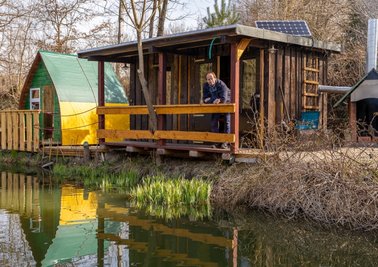 Haustechniker Mirko Buschner an der mit Daniel Glaw erbauten Uferhütte. Links das Kinder-Schlafiglu, rechts der innovative Grill-Kamin. Über einen von der Schlosserei Fleischmann gebauten Wärmetauscher wird Luft eingesaugt und via Rohr in die Hütte zum Heizen geleitet. Die Öko-Immobilie verfügt über einen separaten Zugang und Sonnenplatz.