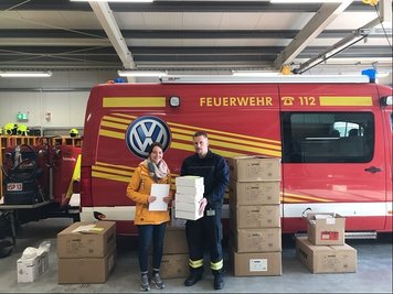 Laura Appelt (l.), Mitarbeiterin der Euro Plus Seniorenbetreuung in Chemnitz, nimmt Atemschutzmasken, Schutzkittel und Schutzbrillen von Werksfeuerwehrmann Thomas Fritzsch auf dem Werksgelände in Zwickau entgegen.