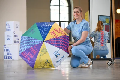 2025-Geschäftsführerin Andrea Pier zeigt den Kulturhauptstadt-Schirm und ein gebatiktes Kosmos-Shirt.