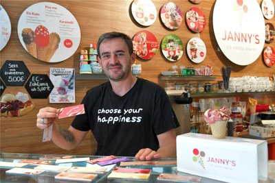 Arben Kosumi steht in „Jannys Eisladen“ und präsentiert ein Sortiment an Eissorten. In der Hand hält er das Schild eines Verkaufsschlagers: Pfirsich Cassis.