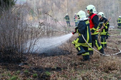 Feuerwehrleute löschen den Vegetationsbrand am Hang nahe einer Gartenanlage.