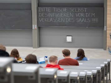            Studierende mit Mund- und Nasenmaske sitzen in einem Hörsaal.