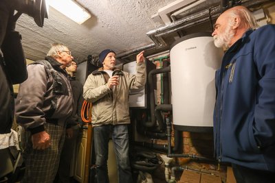 Thomas Grimm (Mitte) hat seinen Keller geöffnet und berichtet von seinen Erfahrungen mit der Wärmepumpe. Sie steht vor dem Haus. Im Keller sind Pufferspeicher und Steuergerät.
