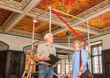 Architekt Uwe Kretzer und Bauamtsleiterin Petra Wolf im Ratssaal im Oederaner Rathaus. Die Deckenbemalung muss ausgebessert werden. Der Erker im Hintergrund wird mit einer Glasscheibe abgetrennt. 