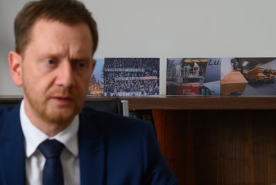 Ministerpräsident von Sachsen sitzt bei einem Interview mit der Deutschen Presse-Agentur in seinem Büro in der Sächsischen Staatskanzlei vor aufgestellten Fotos mit Szenen der Corona-Pandemie. Unter anderen ein Foto des Banners "Kretschmer, du willst Sachse sein? Verhältst dich wie ein Wessischwein!", das beim Zweitligaspiel am 7. November 2021 in Aue von Fans gezeigt wurde.