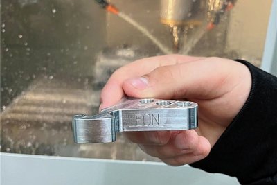Ein Schüler fräste mit einer CNC-Maschine (im Hintergrund) aus einem Metallblock einen als Handyaufsteller verwendbaren Schlüsselanhänger mit Gravur.