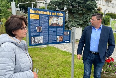 Xenia Brunner und Klingenthals Oberbürgermeister Thomas Hennig an der mit einem QR-Code versehenen Tafel am Marktplatz für den nun mehr-sprachig ausgestatteten Klingenthaler Rundweg. 