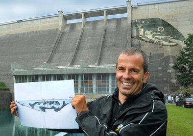 Klaus Dauven zeigt den zeichnerischen Entwurf seines Bildes "Fisch-Reich", von dem ein kleiner Teil schon auf der Mauer zu sehen ist.