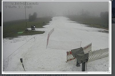 Die Webcam zeigt am Sonntagvormittag eine beschneite Fläche im Nebel. Ringsum ist es grün. 