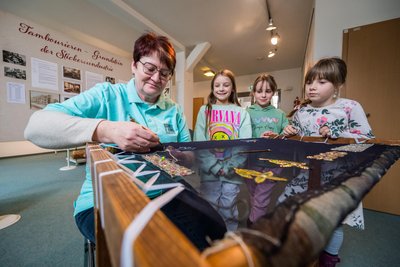 Museumsleiterin Antina Richter hat Charlotte Berisha, Mila Lenk und Kaisa Röder (von links) von der Grundschule Eibenstock die Technik des Tambourierens gezeigt. Die Schule bekommt den Namen Clara-Angermann-Grundschule.