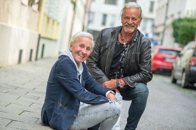 Klaus Eberhartinger (l.), Sänger bei der Ersten Allgemeinen Verunsicherung (EAV), und Thomas Spitzer, Gitarrist und Texter bei EAV, sitzen bei einem Interviewtermin mit dpa auf einem Bürgersteig. Die EAV hört nach über 40 Jahren auf und verabschiedet sich entgegen ihrem humoristischen Ruf mit einem sehr politischen Album.