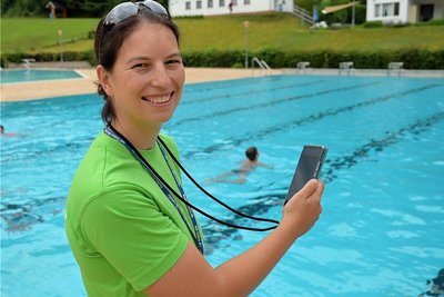 Anne Arnold steht mit der Stoppuhr auf den Handy am Beckenrand und misst die erworbene Ausdauer von Grundschülern beim Schwimmen.