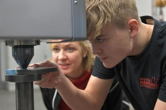 Ausbildungsmarkt Mittelsachsen: Wie Luca seine Lehrstelle fand - Luca Veigl arbeitet am Härteprüfstand seines Ausbildungsbetriebes: Im Hintergrund Sylvia Müller, Mitgeschäftsführerin der G&M Vacutherm Härterei- und Oberflächentechnik GmbH in Brand-Erbisdorf.