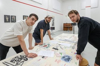 Ausstellungsbesuch: Wie ein Kollektiv den Kapitalismus auseinandernimmt - Béla Bender, Jannis Wolter und Tim Rakutt (von links) von der Gruppe "betrieb.kollektiv" widmen sich derzeit in der Galerie Oscar ganz der "Betriebswirtschaftslehre", wie sie ihre Ausstellung betitelt haben. Zu der gehört auch der große Arbeitstisch in der Mitte des Raums. 