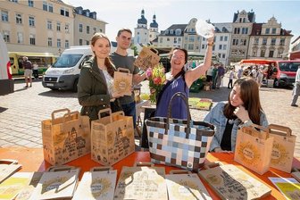 Auszubildende der Plauener Stadtverwaltung kreieren neue Papiertragetaschen für den Handel - Die Auszubildenden Malin Brückner, Frederik Dinter und Pearl Heckel (von links) hatten sich an der Aktion beteiligt. Marktbesucherin Ines Joachim (2. v. r.) freute sich, eine Alternative für Plastik für den Transport ihrer Einkäufe geschenkt zu bekommen.