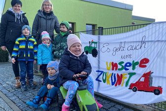 Bauernprotest im Morgenkreis: Was eine Reichenbacher Kita mit einem „kleinen Zeichen“ ausgelöst hat - Mit dem Tret-Traktor und Lynn Könitzer am Steuer auf die Straße. Das Plakat am Zaun der Reichenbacher Kita August Horch hat auch im Internet die Runde gemacht. Hinten im Bild Kita-Leiterin Doreen Seifert (rechts) und Stellvertreterin Anne Hofmann.