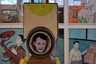 Bilder aus dem beschädigten Leben - Ein "Fasshäuschen" im Vordergrund vor einem bunten Berufe-Bilderbogen von Jan Kummer. 