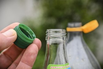 Der Plastikdeckel muss ab - oder doch nicht? - An der Mehrweg-Getränkeflasche lässt sich der Schraubverschluss weiterhin problemlos abnehmen. Anders sieht es bei der Einweg-Flasche im Hintergrund aus. Hier sind Flasche und Deckel seit Juli (fast) unzertrennlich.