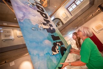 Die Comicfiguren „Vater und Sohn“ feiern in Plauen mit neuer Ausstellung 90. Geburtstag - Interimsgaleristin Sarah Kühnel und Martina Albig hängen das Raumbanner auf, welches auf die Geburtstagsausstellung hinweist.