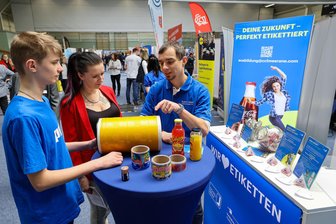 „Die Geld-Frage wird nicht mehr zuerst gestellt“: Wie sich die VW-Krise auf Ausbildungsmarkt im Landkreis Zwickau auswirkt - Sie hielten am Stand von CCL Label: Der 14-jährige Sidney (mit seiner Mutter Stefanie) sprechen mit Druck-Medientechnologe Robert Veiland bei der Ausbildungsmesse in Meerane.