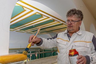 Ein Goldband ziert wieder den Ratssaal im Falkensteiner Rathaus - Malermeister Axel Richter streicht an der Empore über dem Ratssaal ein Stück Goldband nach. Hinter ihm die Decke des Ratssaals. Zuletzt waren dort blaue Felder mit roten Rahmen, jetzt sind Grün und Gold zurück.