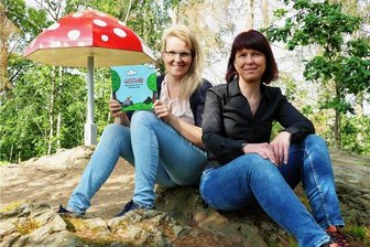 Ein Kinderbuch als Jubiläumsgeschenk: Wie Gelenau vor 750 Jahren besiedelt wurde - Am Gelenauer Fliegenpilz, wo die Waldwichtel Moosbert und Waldfried zuhause sind, präsentieren Kristina Schreckenbach (links) und Christine Drechsel ihr Kinderbuch „Gelenau - Ein neues Dorf im Finsterwald“.