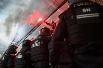 Ein leichtverletzter Beamter, ein am Kopf verletzter Fan, ein Angriff mit Pyrotechnik: So lief das Risikospiel zwischen Erzgebirge Aue und Energie Cottbus - Polizisten stehen im Erzgebirgsstadion in Aue vor dem Cottbuser Fanblock, während ein Fan auf dem Zaun sitzt und Pyrotechnik zündet.