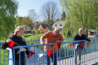 Ersterwähnung als säumiger Schuldner: St. Egidien feiert „700fünf“ – obwohl es schon mal 1000 Jahre alt war - Julia Redlich vom Gemeindeamt sowie André Schatz und Gerhard Sonntag vom Festkomitee (v. l.) probieren an der Rathaus-Brücke schon mal eine Wimpelkette aus.