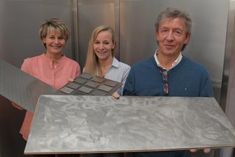 Volker Queck (rechts) zeigt die allererste Edelstahl-Bodenplatte, mit der er zu experimentieren anfing. Seine Frau Katrin (links) hält eines der inzwischen patentierten Paneele, Tochter Josefine präsentiert die zwölf derzeit verfügbaren Oberflächen-Varianten. Im Hintergrund eine Wand aus Queck-Edelstahl-Segmenten.